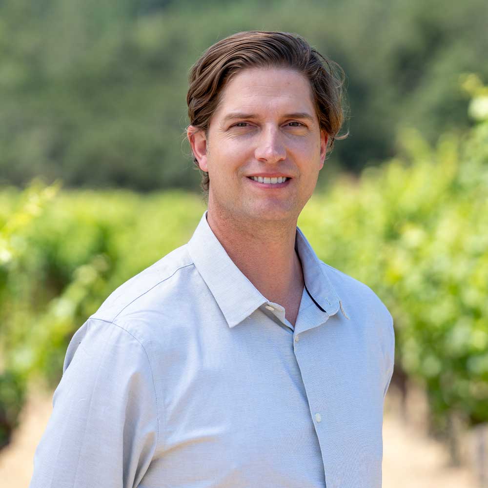 Man standing in a vineyard wearing a light-colored shirt.