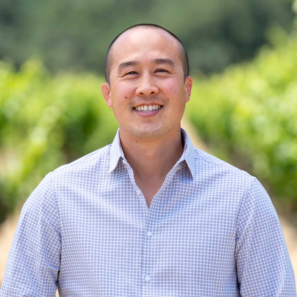 Man wearing a light blue checkered shirt outdoors with greenery in the background