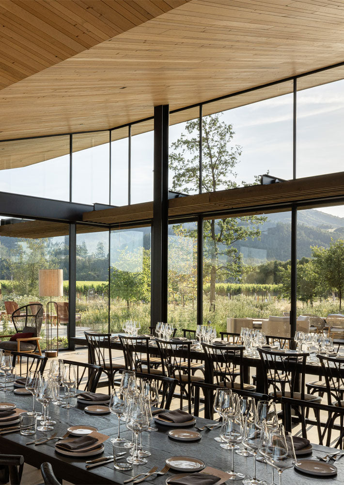 A large, modern event space with high wood-paneled ceilings, floor-to-ceiling windows, and black dining tables and chairs set with glassware and plates, overlooking a vineyard and mountains.